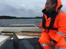 Mann i oransje kjeledress og redningsvest sittende med en bærbar datamaskin i en småbåt ute på fjord med land i bakgrunnen.