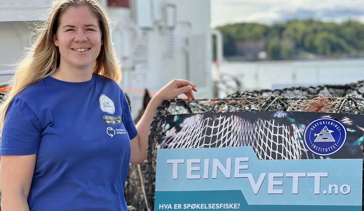 
Kvinne smiler til kamera, står foran plakat 